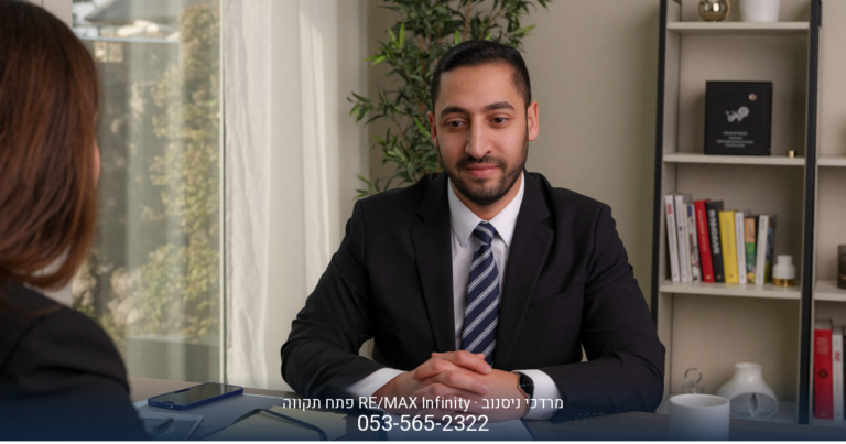 מרדכי ניסנוב, מתווך RE/MAX בפתח תקווה, יושב בחליפה מול מדף ספרים ועציץ